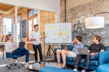 Photo by Austin Distel three men sitting while using laptops and watching man beside whiteboard